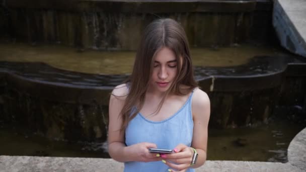 Eine junge Frau sitzt auf einem Stadtbrunnen und benutzt ein Mobiltelefon. Mobile Apps, mobiles Internet — Stockvideo