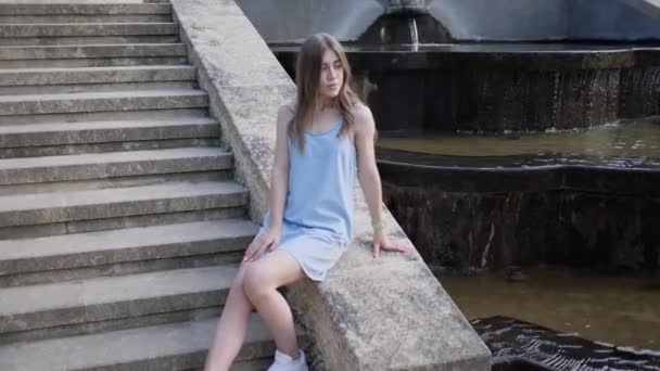 Tânără femeie frumoasă în rochie stătea jos pe scări lângă fântâna orașului — Videoclip de stoc