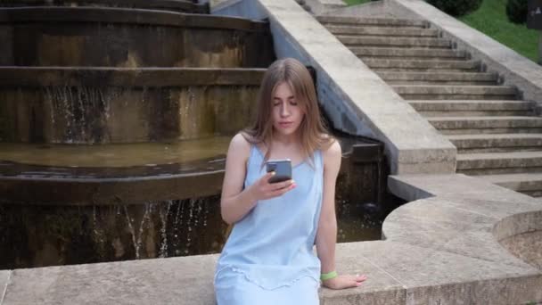 Junge schöne Frau sitzt auf Stadtbrunnen in der Stadt und benutzt Handy. Konzept der telefonabhängigen Menschen — Stockvideo