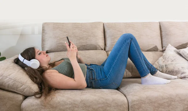 Девочка-подросток лежит на диване дома и смотрит в телефон. — стоковое фото
