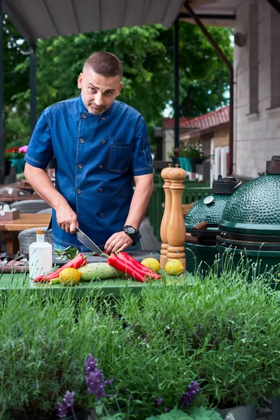 Hermoso chef de aspecto europeo de pie en la terraza del restaurante, cortando verduras y mirando a la cámara — Foto de Stock