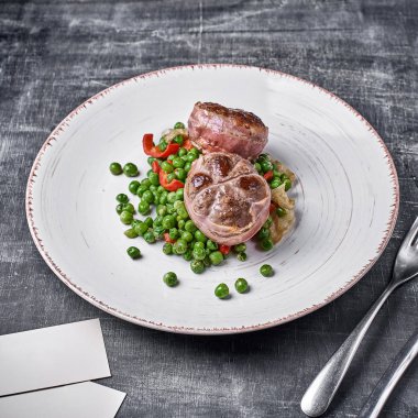 Dana madalyon içinde pastırma sarılmış, yeşil bezelye ile ve ahşap arka plan üzerinde bir plaka üzerinde hizmet vermektedir. Yakın, seçici odak. 1:1