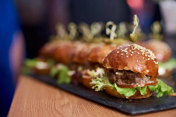 Soyut bulanık gıda arka plan. Et, salata peynir ve domates açık havada yaz barbekü partisi sırasında ahşap plaka ile küçük hamburger. Gıda hazırlama, sohbet ve içki yiyorsunuz arkadaşlar. Yakın çekim