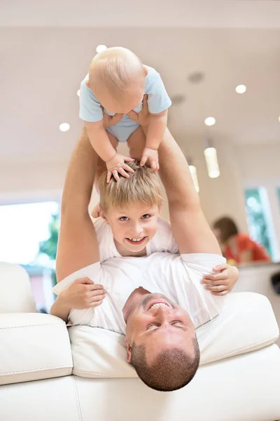 Concepto familiar, padre jugando en el sofá con el bebé y su hermano, infancia feliz. Un disparo interior en la cocina. un bebé en sus brazos, un bebé arroja al techo, familia feliz — Foto de Stock