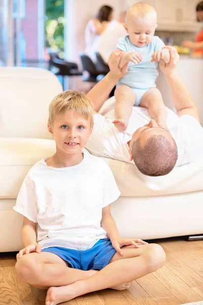 O conceito de família, o menino senta-se no chão olhando para a câmera, próximo pai brinca no sofá com o bebê, a criança senta-se no peito do pai, infância feliz, família — Fotografia de Stock