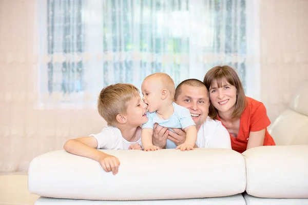 Concepto familiar, padres jóvenes con dos hijos, el bebé muerde la nariz de su hermano. Una infancia divertida y feliz. Familia en el sofá mirando a la cámara, sonriendo. una familia feliz — Foto de Stock