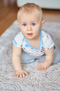 Büyük mavi gözlü sevimli gülümseyen küçük çocuk kameraya bakıyor, dört parça üzerinde sürünerek.