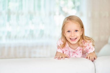 Mutlu sevimli küçük Blonde kız, evde odada beyaz kanepe arkasında gizlemek, portre . Kameraya mutlu bir şekilde bakmak.
