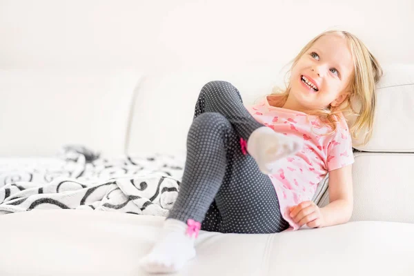 Happy cute little Blonde girl, hide behind the white sofa in the room at home, portrait. Смотреть счастливо в камеру . — стоковое фото