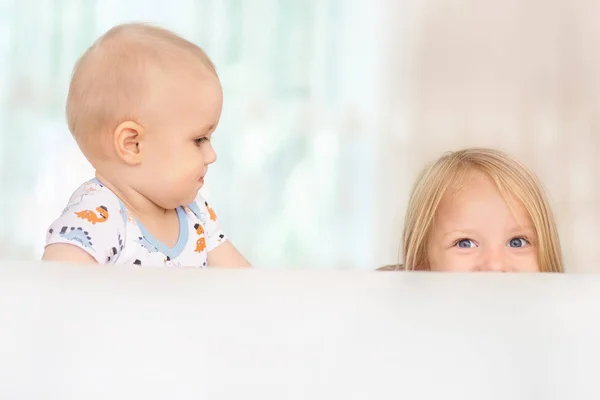 Dos dulces niños caucásicos felices hermanos niña y niño se esconden detrás del sofá blanco en la habitación en casa, retrato. Mirando felizmente a la cámara, en la luz amplia sala de estar, estar solo en casa . —  Fotos de Stock