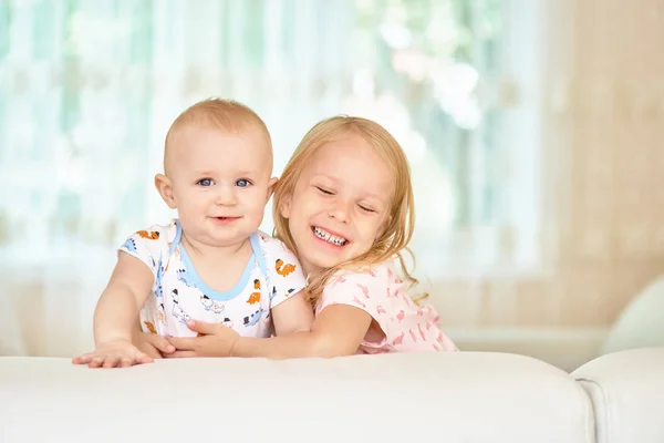 Two sweet happy Caucasian children siblings girl and boy hide behind the white sofa in the room at home, portrait . Looking happily at the camera, in light spacious living room, being home alone. Stock Image