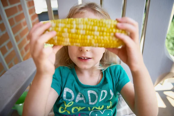 Schattig meisje blonde met maïs in haar handen, zonnige dag, op het balkon van het huis, het groen op de achtergrond. Agrarische oogst, maïs — Stockfoto