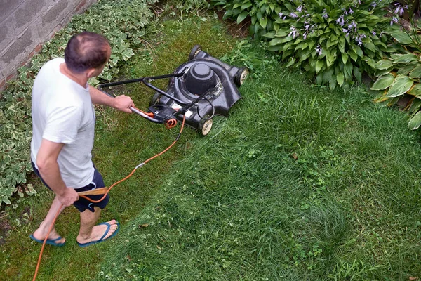 Middelbare leeftijd man in shorts en t-shirt, snijden begroeid gras met ouderwetse grasmaaier in suburbane huis — Stockfoto