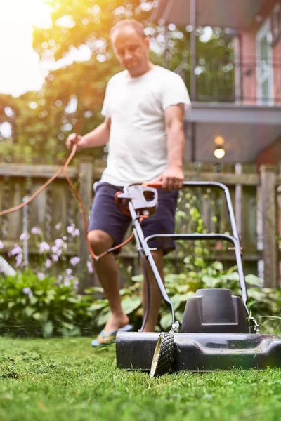 Middelbare leeftijd man in shorts en t-shirt, snijden begroeid gras met ouderwetse grasmaaier in suburbane huis — Stockfoto