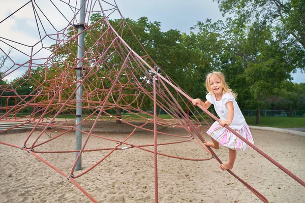 Маленькая девочка улыбается, смотрит в камеру, маленький ребенок играет в лазание по сети. на открытом воздухе в солнечный летний день . — стоковое фото