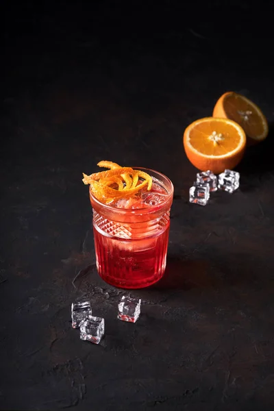 Negroni-Cocktail mit orangefarbenem Twist and Pin, auf dunklem Hintergrund, daneben Orange und Eis. Getränkefotografie. — Stockfoto