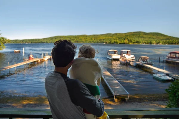 Young man with a little girl are standing on the veranda of the house and looking at the lake. Early morning, sunny day. Early cold morning in the fall.