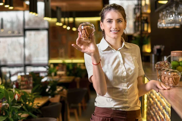 Positive young waitress suggests, stretching a little box of cookies in a sweets shop. Copy space for text — Stock Photo, Image