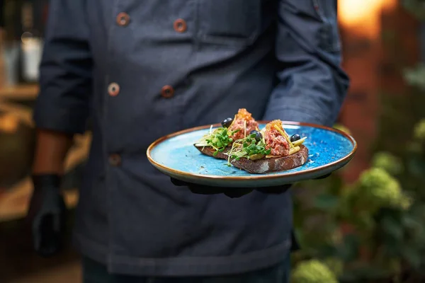 Leckeres rohes Rindfleisch-Tatar auf Bruschette, das Gericht hält ein Koch in blauer Uniform in der Hand. — Stockfoto