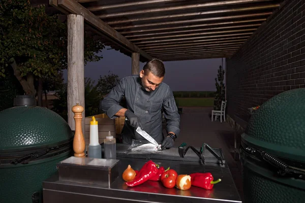 El chef cocina caballa de pescado fresco, espolvorea con sal marina y verduras. Foto horizontal. Concepto de preparar cocina saludable y vegana, una terraza nocturna de un restaurante, negocio hotelero. Tecla baja — Foto de Stock