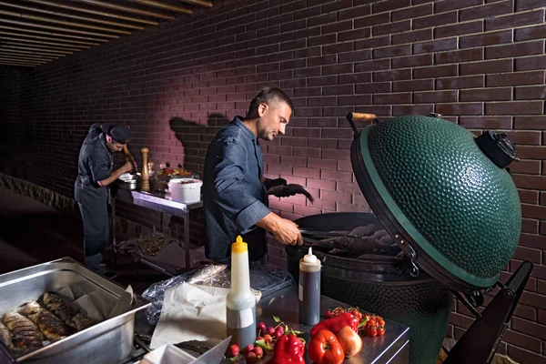 Chef y asistente, cocinar caballa fresca, espolvorear con sal marina y verduras. Foto horizontal. Concepto de preparación de cocina saludable y vegana, una terraza nocturna de un restauranteLow key — Foto de Stock