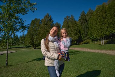 güneşli bir günde parkta yürüyorlar gibi yürümeye başlayan çocuk kızı kucağında tutan genç sarışın anne