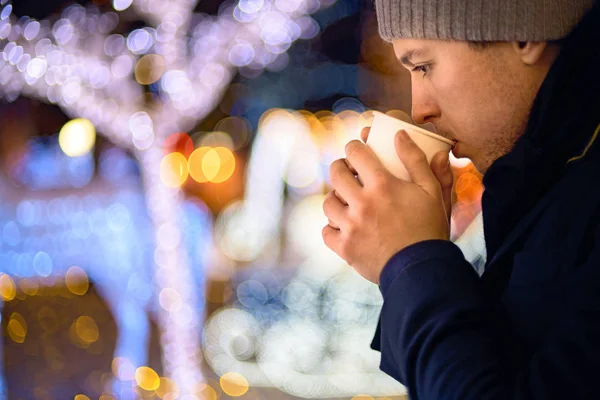 The guy holds holds hot tea in his hands on a background of Christmas lights. hot drinks, holidays and people concept - close up