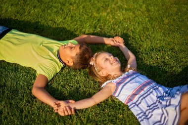 Yeşil çimlerin üzerinde uzanmış gülümseyen iki çocuğun portresi. Neşeli erkek ve kız kardeş birlikte gülüyorlar. Mutlu çocuklar park çimlerinde eğleniyorlar. Çocuk günü