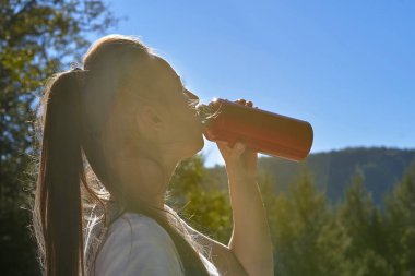 Beyaz tişörtlü çekici bir sarışın şişeden su içiyor, güneşli bir yaz gününde dağlarda duruyor. Rötuş, parlak renkler
