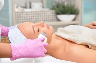 Yakın plan kozmetolojist, genç ve güzel bir kadının yüzünün ultrason donanımını temizliyor. Cilt temizleme ve esnekliğin yenilenmesi kavramını benimse