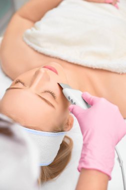 Yakın plan kozmetolojist, genç ve güzel bir kadının yüzünün ultrason donanımını temizliyor. Cilt temizleme ve esnekliğin yenilenmesi kavramını benimse