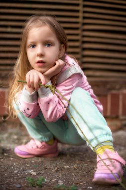 Oturan kız çömelmiş kameraya bakıyor. Pembe kazaklı 6 yaşında bir kız. Kızın uzun sarı saçları var. Seçici odak resmi.