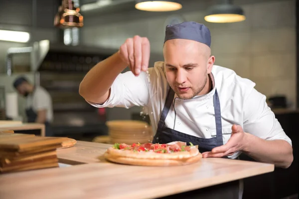 Food concept. Preparing traditional italian pizza. Young smiling chef in white uniform and gray hat decorate ready dish in interior of modern restaurant kitchen. Ready to eat — Stock Photo, Image