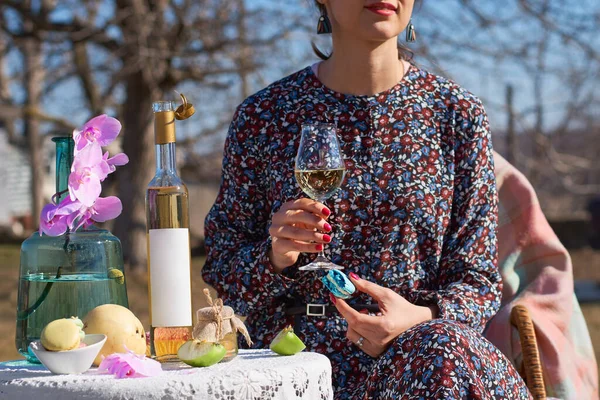Открытый стол с тарелкой закусок и бутылкой с пустым этикетом для макета. Девушка с бокалом вина в руках в солнечный день . — стоковое фото