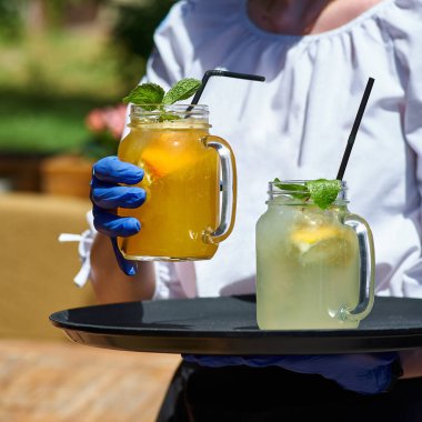 Garsonun elinde turuncu ve limonlu mason kavanozlarda soğuk ve ferahlatıcı limonata. Yaz terasında güneşli bir gün.