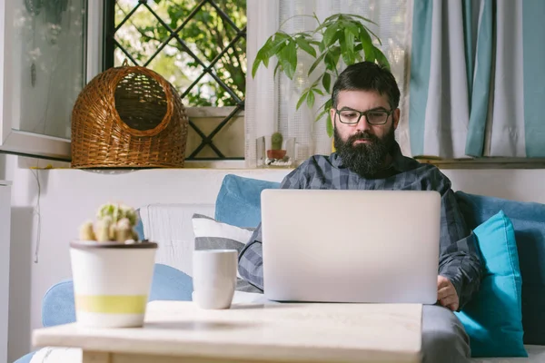 bearded young man working with laptop while sitting on sofa