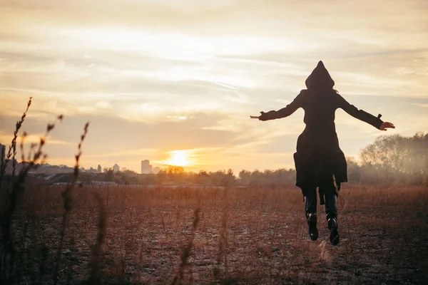 back view of young woman in black coat with hood jumping with open arms on field at sunset