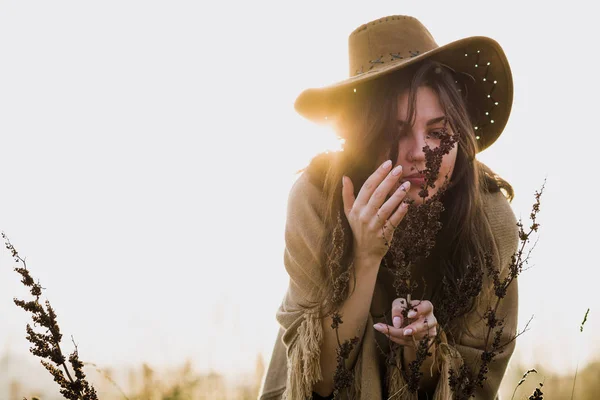 beautiful brunette cowgirl in hat touching dry plants on meadow at sunset