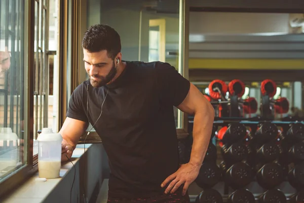 sportive man standing by gym window with protein shake and using smartphone with earphones