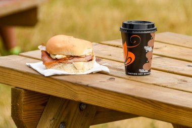 Fairford, İngiltere - Temmuz 2018: Sıcak domuz pastırması rulo ve bir bir tek kullanımlık plastik bardak kahve ahşap bir piknik masa açık bir olay üzerinde.