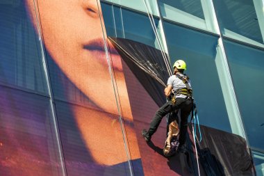 CANNES, FRANCE - APRIL 2019: Specialist maintenance operative abseiling down the side of the Palais des Festivals in Cannes to remove plastic advertising hoarding from the glass panels of the building. clipart