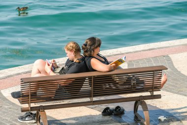 Lazise, Lake Garda, İtalya - Eylül 2018: Garda Gölü'ndeki Lazise'deki gezinti yolundaki ahşap bankta dinlenen iki kişi.