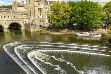 Bath, İngiltere - Temmuz 2019: Avon Nehri üzerindeki pulteney Weir ve Bath'ın merkezinde Pulteney Köprüsü şeklinde. Bir turist teknesi arka planda.