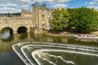 Bath, İngiltere - Temmuz 2019: Bath'ın merkezinde Avon Nehri ve Pulteney Köprüsü üzerinde at nalı şeklindeki Pulteney Weir'ın geniş açı görünümü. Bir turist teknesi arka planda.