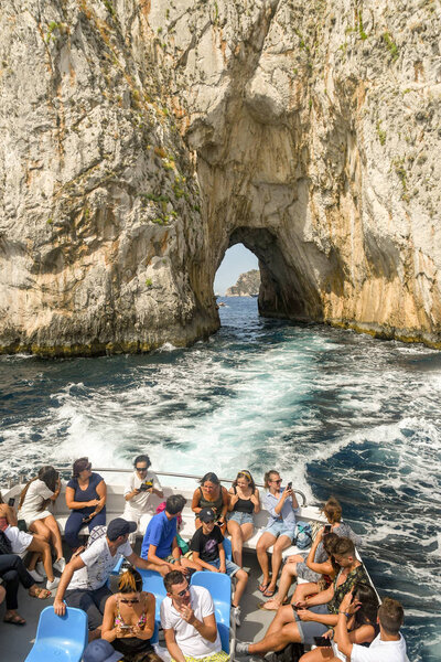 Остров Каприи, Италия - АВГУСТА 2019: Люди на заднем сиденье лодки, которая прошла через арку в скале Фаральони у берегов Капри
.