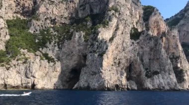 CAPRI ISLE, ITALY - 2019 AĞUSTOS: Capri Adası 'nın engebeli kıyı şeridi. Uçurumun önünde küçük bir tekne hareket ediyor..
