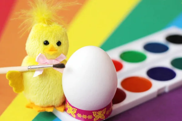 Happy Easter. Yellow toy chicken with a brush is preparing to paint a white chicken egg watercolor.