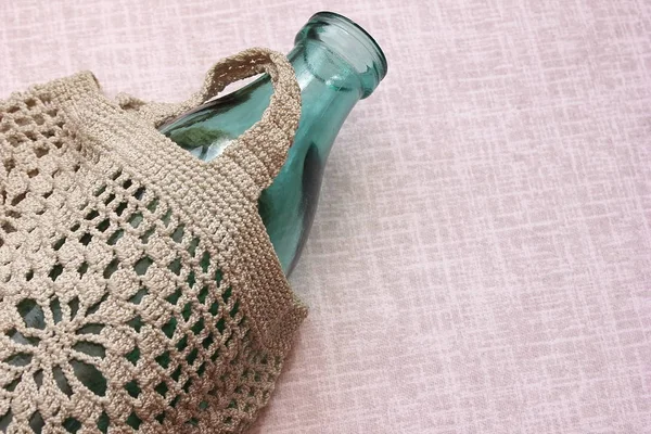 The glass bottle is in a knitted bag on the linen tablecloth. Reusable packaging. — Stock Photo, Image