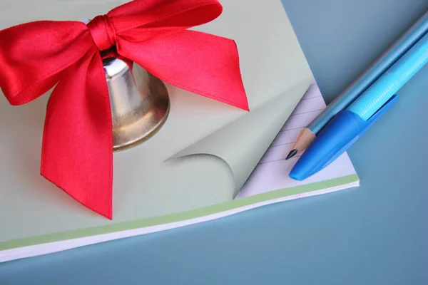 Eine Metallglocke mit roter Schleife steht neben Stift und Bleistift auf dem Schulheft. — Stockfoto