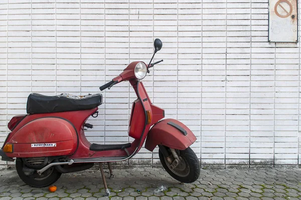 Old Vespa-like red motorbike parked in front of a grid white wall.